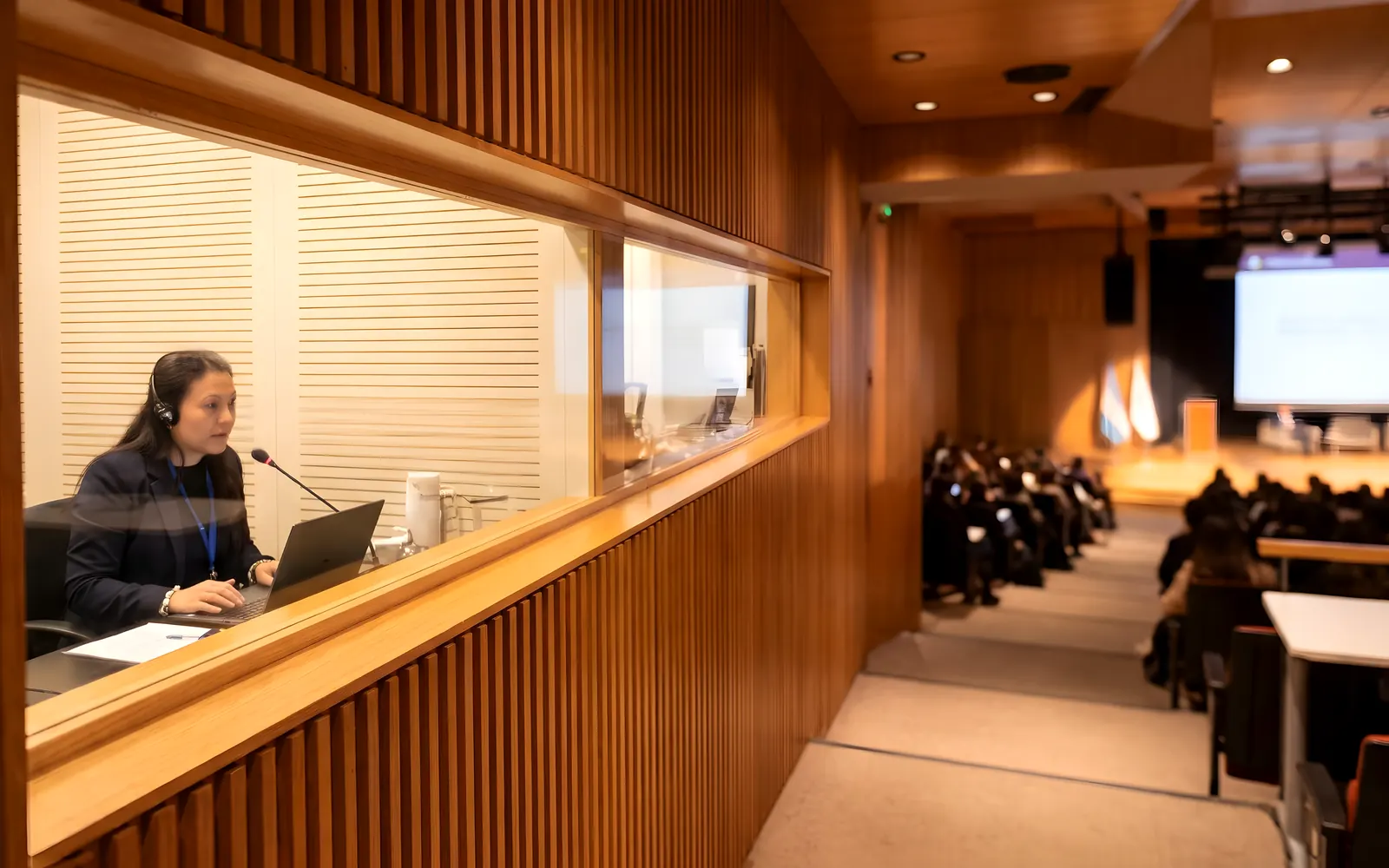 Intérprete con auriculares trabajando en la cabina de traducción simultánea, con el auditorio y el escenario al fondo